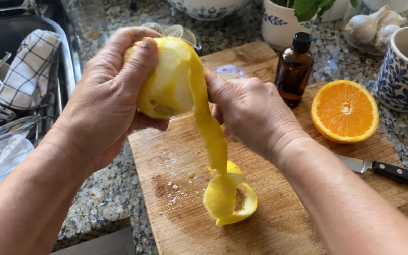 Hands peeling a lemon next to an orange half and a small amber essential oil bottle on a cutting board